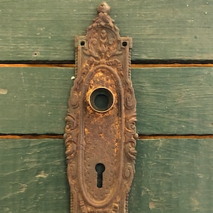 May include: Ornate, antique door plate with a keyhole and a round knob opening. The metal plate has a weathered, rusty appearance with decorative floral and scrollwork details. The background is a green wooden door.