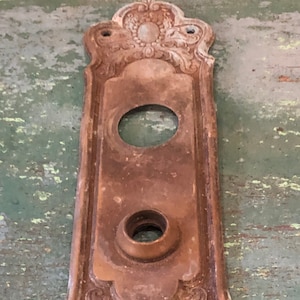 May include: Antique brass door plate with ornate floral design and two keyholes. The plate has a worn, aged patina.