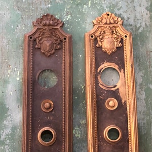 May include: Two antique brass door plates with ornate gold trim and a shield design. Each plate has three round buttons.