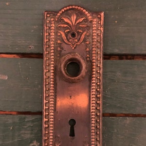 May include: Antique copper door plate with a keyhole and ornate design. The plate has a beaded border and a floral pattern.