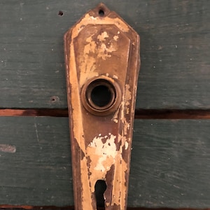 May include: Vintage brass door plate with a keyhole and a circular opening for the doorknob. The plate has a distressed finish with peeling paint, revealing the aged metal underneath. The plate is mounted on a dark green wooden surface.