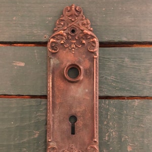 May include: Antique brass door plate with ornate detailing and a keyhole. The plate has a worn, aged look.