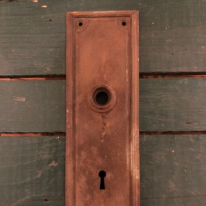 May include: A rectangular, rusty metal door plate with a keyhole and a round knob. The plate has a decorative border and is mounted on a green wooden surface.