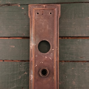 May include: A vintage brass door plate with two keyholes and a decorative design. The plate is a dark brown color with a patina finish.