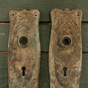 May include: Two antique brass door plates with ornate designs and keyholes. The plates are slightly tarnished and have a greenish patina.