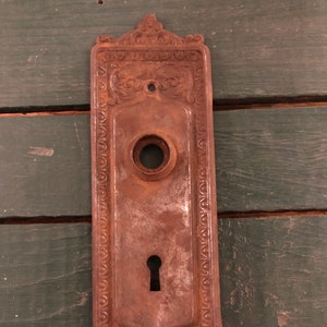 May include: A rusty, ornate, metal door plate with a keyhole and a round knob. The plate has a decorative border and intricate floral designs.