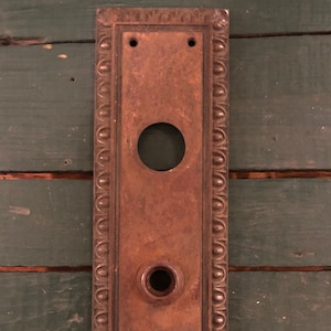 May include: A vintage, rectangular, rusty metal door plate with an ornate border and two keyholes. The plate has a circular cutout in the center.