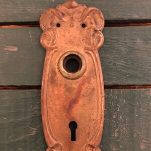 May include: An antique brass door plate with an ornate design and a keyhole. The plate is slightly tarnished and has a rustic look.