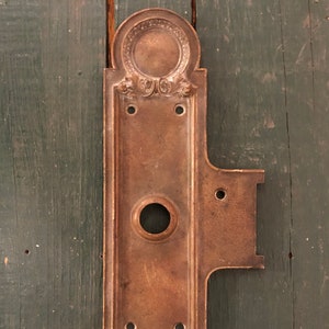 May include: Antique brass door plate with ornate details and a keyhole. The plate has a rectangular shape with rounded edges and a decorative design.