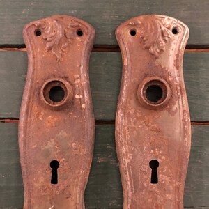 May include: Two antique rusty metal door plates with ornate designs and keyholes. The plates are a dark brown color with a patina of rust.