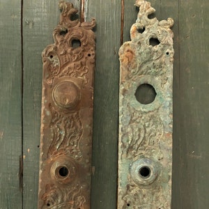 May include: Two antique door plates with ornate designs and a worn, aged patina. The plates are made of metal and have a dark brown and green color.