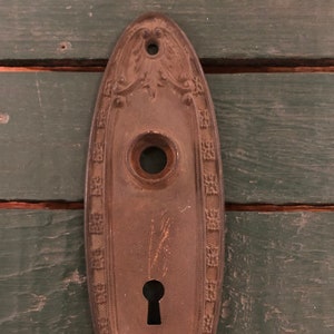 May include: Antique oval-shaped door plate with ornate detailing, a keyhole, and a round knob. The plate is made of metal and has a brown patina.