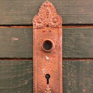 May include: Antique brass door plate with ornate floral designs and a keyhole. The plate has a worn, aged look.