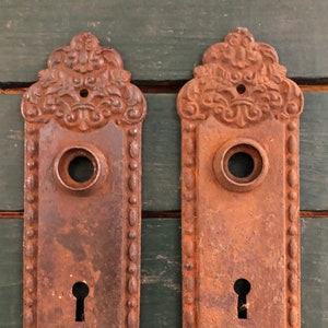 May include: Two antique, rusty, cast iron door plates with ornate designs. Each plate has a keyhole and a round hole for a knob.
