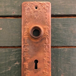 May include: A rusty metal door plate with an ornate floral design. The plate has a keyhole and a round hole for a doorknob.