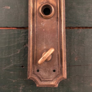 May include: A vintage brass door plate with a keyhole and a decorative knob. The plate has a rectangular shape with a decorative border and is slightly tarnished.