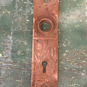 May include: A vintage brass door plate with an intricate design featuring a keyhole and a circular cutout. The plate has a worn, aged patina.