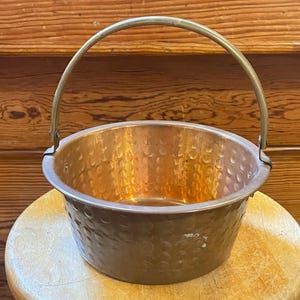 May include: A copper cauldron with a hammered texture and a brass handle. The round pot sits on a wooden stool, with the handle arched above the rim. The copper has a warm, reddish-brown hue, reflecting light.