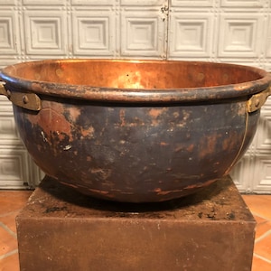 May include: A large, antique copper cauldron with a dark patina. The bowl-shaped pot has two sturdy handles and sits on a dark, square base. The interior of the cauldron is a bright copper color, contrasting with the darker exterior.