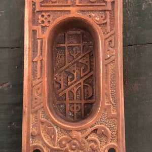 May include: A vintage, ornate, copper-toned door or window pull with an oval-shaped opening and intricate floral and geometric designs.