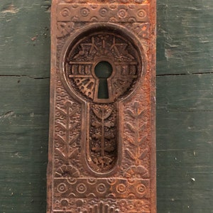 May include: A vintage, ornate, cast iron door plate with a keyhole. The plate is rusty brown and has a decorative design of circles, leaves, and geometric shapes.