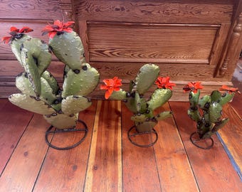 May include: Three metal cactus sculptures with red flowers. The sculptures are on a wooden surface.