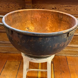 May include: A large copper pot with two handles, showing signs of wear and age. The pot is sitting on a white stool, with a wooden floor in the background.