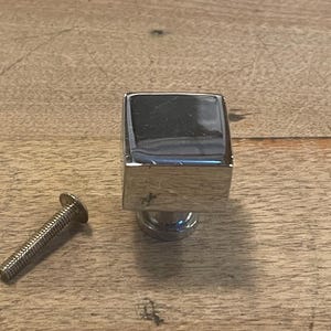 May include: A chrome-finished square cabinet knob with a matching screw. The knob has a reflective surface and a simple, modern design. The hardware is shown on a wooden surface.