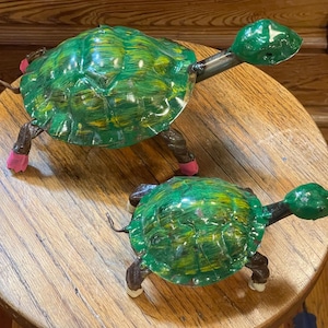 May include: Two handmade turtle sculptures with green and yellow shells. The turtles have metal necks and legs, with pink accents on their feet. They are displayed on a wooden surface.