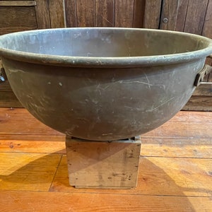 May include: A large, round, copper bowl with two handles. The bowl is sitting on a wooden block.