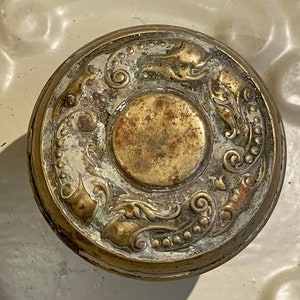 May include: A vintage brass door knob with an ornate design featuring a central circle surrounded by swirling floral patterns.