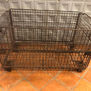 May include: A large, rusty metal wire storage crate with wheels. The crate is made of a wire mesh and has two levels. The crate is sitting on a tiled floor.