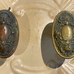 May include: Two oval-shaped antique brass door knobs with intricate floral designs. The knobs have a worn, aged patina and are a beautiful example of vintage hardware.