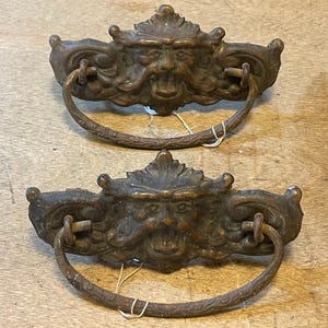 May include: Two matching bronze-toned metal drawer pulls featuring a central relief of a bearded face with ornate scrollwork detailing. Each pull has a wide, curved, hinged handle attached to the decorative backplate with two metal loops.