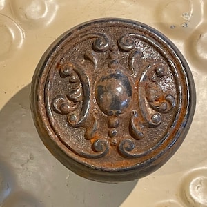 May include: A close-up of a round, ornate, cast iron knob with a detailed floral design. The knob is rusty brown and has a raised center.