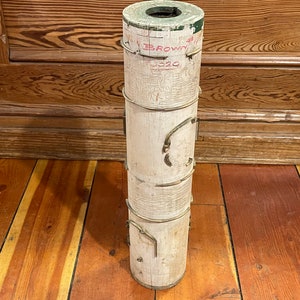 May include: A white wooden cylinder with green and red painted text that reads "S. Brown 320". The cylinder has several brass rings and is sitting on a wooden floor.