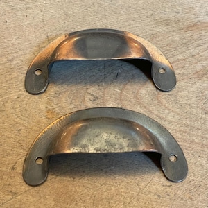 May include: Two antique-style, half-moon drawer pulls with a copper-colored finish. Each pull has two mounting holes and a slightly distressed appearance, suggesting age or vintage style. The pulls are made of metal and are set against a wooden surface.