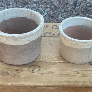 May include: Two cylindrical planters. The larger one is a muted brown with a white band near the top. The smaller planter has a similar design. Both are sitting on a wooden surface.