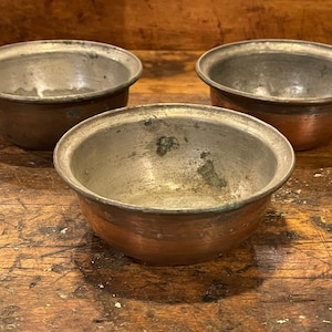 May include: Three antique copper bowls with a tarnished silver interior. The bowls are sitting on a wooden surface.