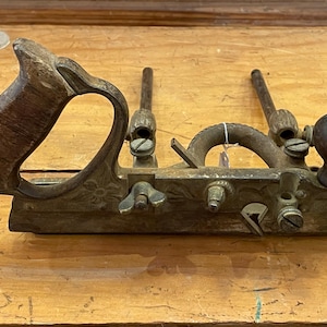 May include: An antique woodworking tool, a hand plane, rests on a wooden surface. The plane features a wooden handle and knob, with metal components showing signs of age. The design includes decorative floral patterns and adjustable parts.
