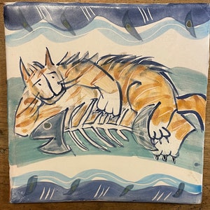May include: A hand-painted ceramic tile featuring a cartoon illustration of an orange cat with black stripes lying on top of a fish skeleton. The background is a blue and white watercolor design with wavy lines and small blue fish.