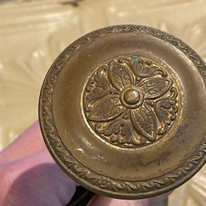 May include: A gold-toned metal decorative knob with an ornate floral design. The knob has a raised center and a detailed border.