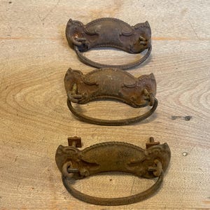 May include: Three antique bronze drawer pulls with ornate, curved designs. Each pull features a decorative face and a curved handle. The pulls are arranged vertically against a light brown wooden surface, showcasing their aged, rustic appearance.