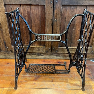 May include: A black cast iron Singer sewing machine base featuring an ornate, curved frame with a central crossbar displaying the word SINGER in raised silver lettering and a rectangular foot pedal with a decorative lattice pattern at the bottom.