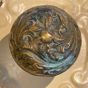 May include: A brass door knob with an ornate, floral design. The knob has a greenish patina.