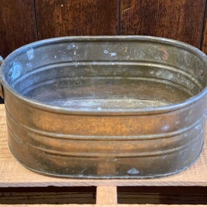 May include: An oval-shaped, copper planter with two handles. The planter has a worn, aged look and is made of metal.