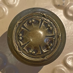 May include: A brass door knob with a decorative, ornate design. The knob has a circular shape with a raised, cross-shaped center.