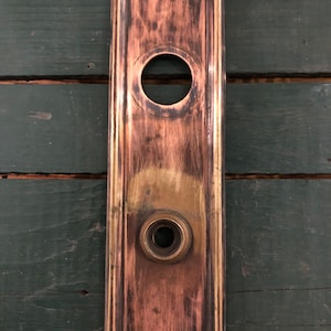 May include: Antique brass door plate with a rectangular shape and two keyholes. The plate has a worn, aged finish with a copper patina.
