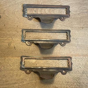 May include: Three antique brass drawer pulls with rectangular label holders. Each pull has a decorative handle and mounting holes. The brass has a weathered patina, suggesting age and use. The pulls are arranged on a wooden surface.