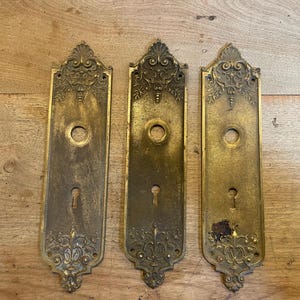 May include: Three antique brass door plates with ornate floral and scroll designs. Each plate has a keyhole and a circular opening for the doorknob. The plates are rectangular with a decorative top and bottom, showing signs of age and wear.
